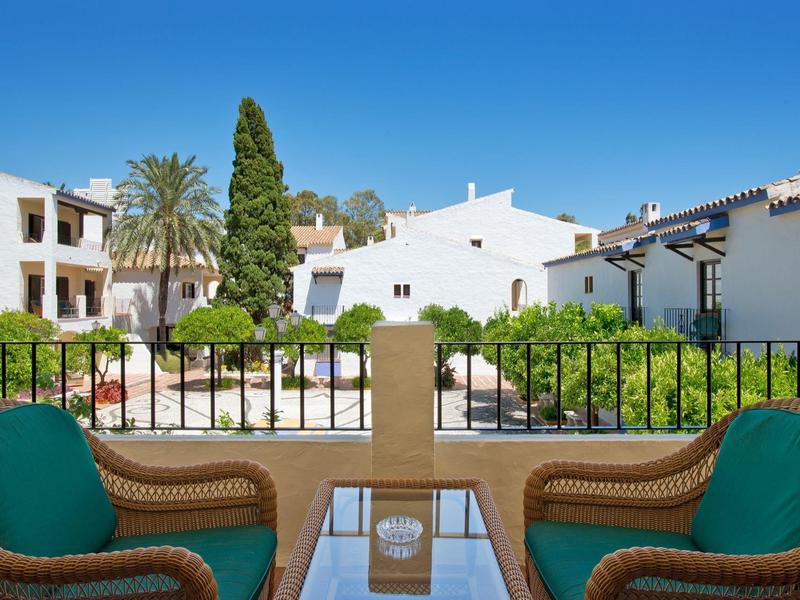 Vue sur une terrasse calme avec deux chaises et des coussins verts, des bâtiments blancs et des plantes au-delà.