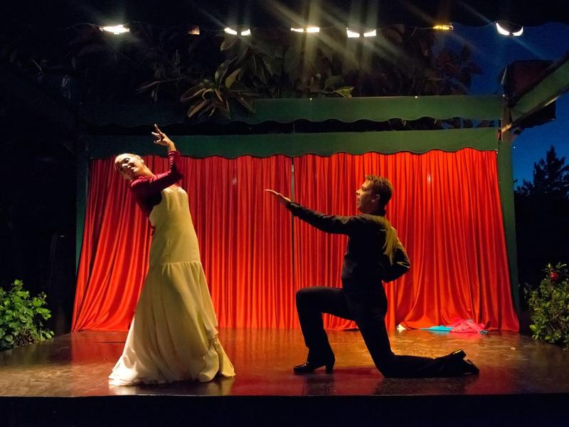 Un couple danse sur une scène avec un rideau rouge et un fond sombre.