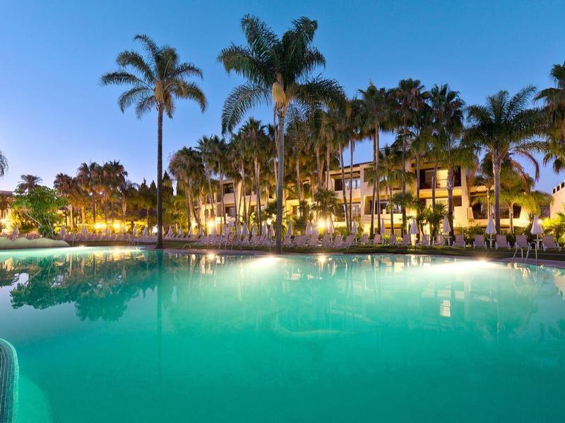 Grand piscine éclairée avec palmiers et chaises longues dans un hôtel tropical la nuit.