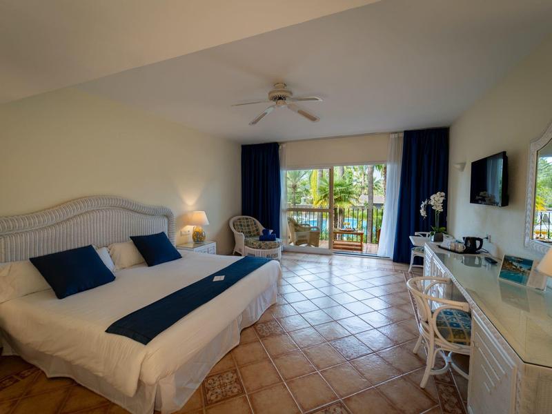 Chambre d'hôtel lumineuse avec grand lit, TV à écran plat, bureau, ventilateur de plafond et balcon avec vue sur la verdure.