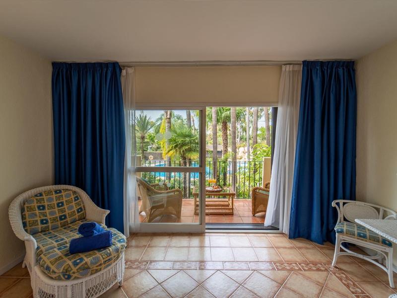 Chambre lumineuse avec fauteuil en osier, table et vue sur balcon avec palmiers et piscine.