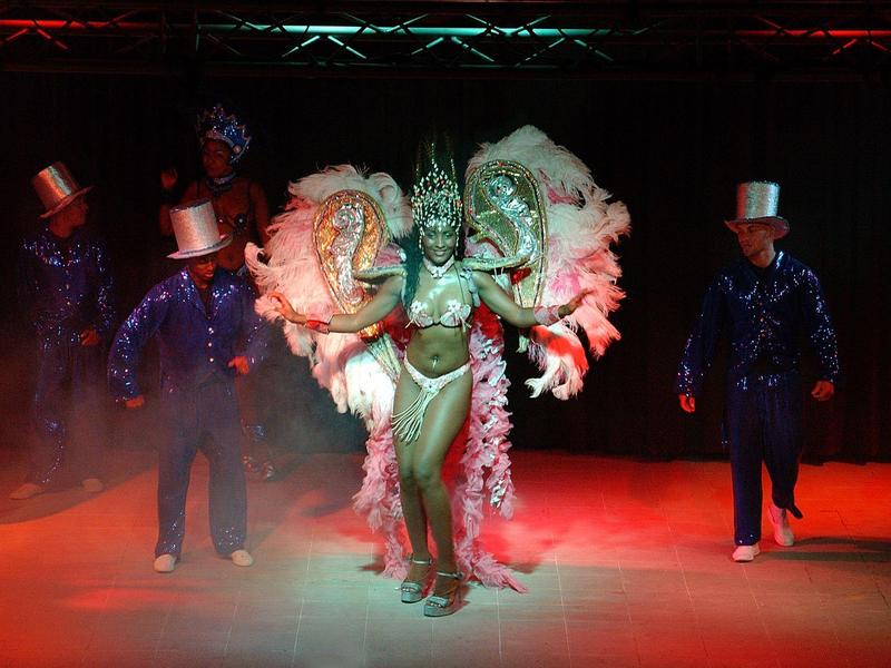 Dancer in colorful feather costume with two companions in glitter suits and top hats.