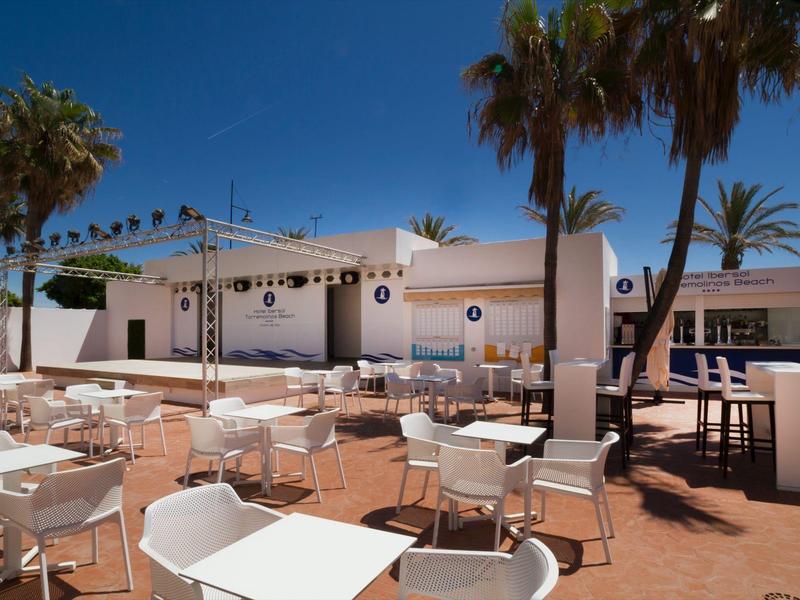 Sunny hotel terrace with white tables and chairs under palm trees.