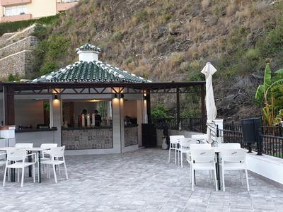 Area esterna dell'hotel con tavoli, sedie e un bar coperto di fronte a una collina boscosa.