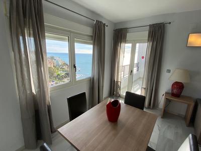 Stanza luminosa con tavolo in legno, vaso rosso e vista sul mare attraverso finestra e porta del balcone.