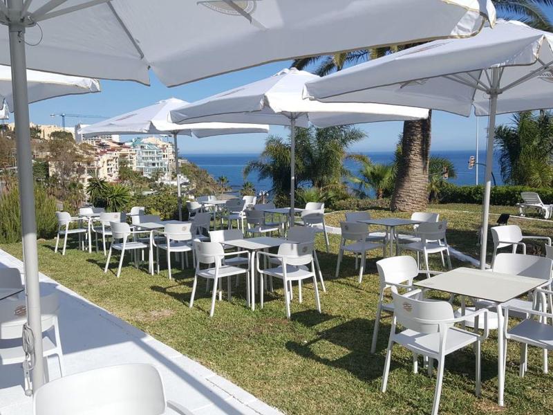 Area esterna dell'hotel con tavoli e sedie bianchi sull'erba sotto ombrelloni con vista sul mare.