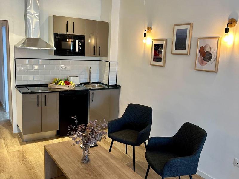 Area cucina moderna con pavimento in legno, unità cucina nera e due poltrone blu vicino a un tavolo in legno.