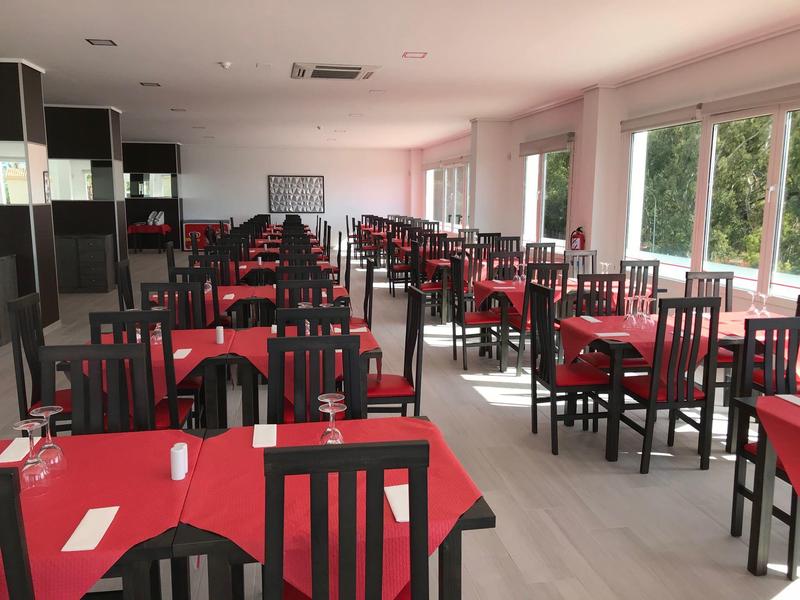 Ampia sala da pranzo con tavoli e sedie, tovaglie rosse e bicchieri apparecchiati.