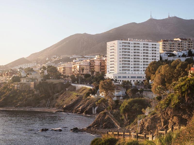 Città costiera con spiaggia, edifici di hotel sulla collina e montagne sullo sfondo al tramonto.