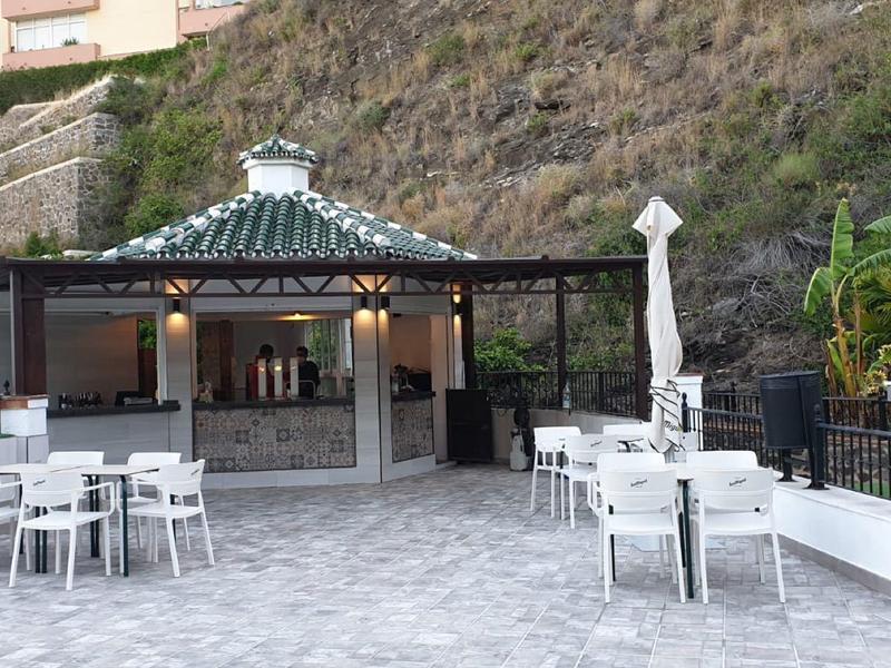 Area esterna dell'hotel con tavoli, sedie e un bar coperto di fronte a una collina boscosa.