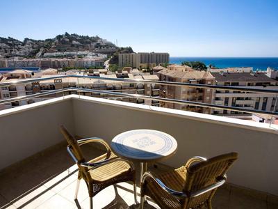 Balcon avec table et chaises offrant une vue sur la ville, les collines et la mer sous un ciel clair.
