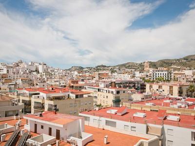 Paysage urbain avec des toits rouges et des montagnes en arrière-plan sous un ciel nuageux.