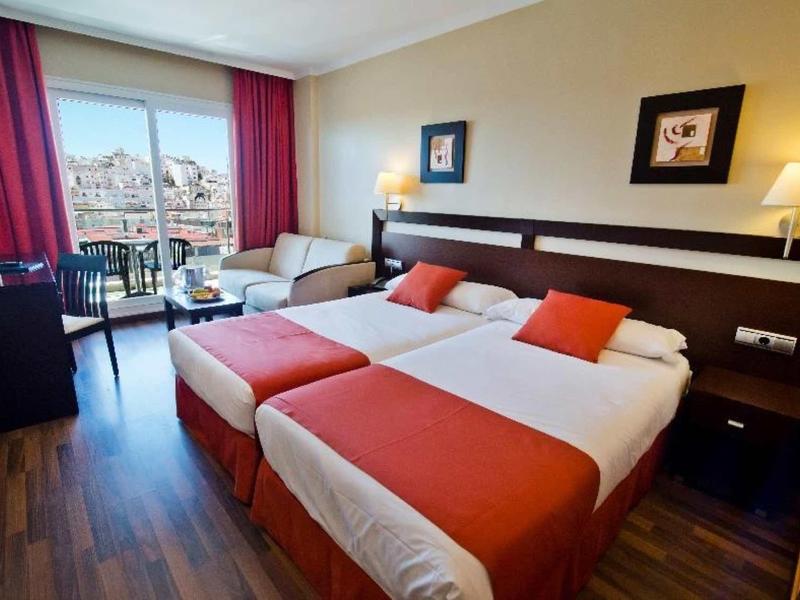 Chambre d'hôtel avec deux lits simples, accents rouges et vue sur la ville à travers de grandes fenêtres.