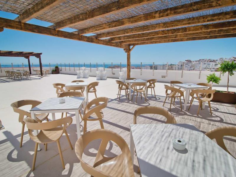 Terrasse couverte avec tables et chaises, vue sur la ville et la mer en arrière-plan.