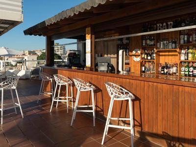 Outdoor hotel bar with wooden counters, white stools, and a view of poolside seating.