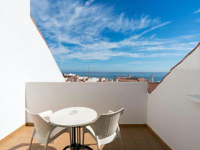 Small balcony with two white chairs and table overlooking buildings and sea under blue sky.