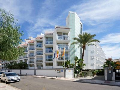 Modern white hotel building with balconies, palm tree, and cars parked nearby under blue sky.