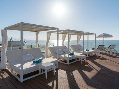 White canopy beds and lounge chairs on a wooden deck by the sea under a bright sun.