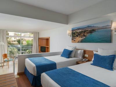 Hotel room with two beds, blue and white bedding, sea view painting, and a balcony with chairs.