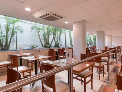 Spacious hotel dining area with wooden chairs and tables, large windows with greenery outside.