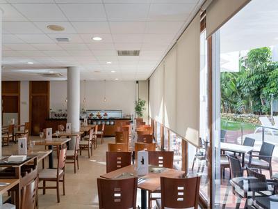 Bright restaurant with wooden chairs and tables next to large windows with blinds