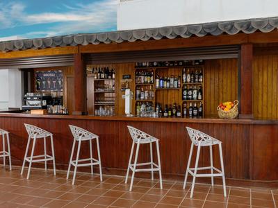 Bar mit Holztresen und weißen Barhockern vor einem Fenster mit blauen Himmel.