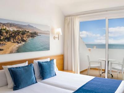 Helles Hotelzimmer mit zwei Betten, blauen Kissen und Meerblick durch die Glasfront.
