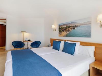 Modernes Hotelzimmer mit Doppelbett, blauer Bettdecke, zwei Sesseln und Meeresbild an der Wand.