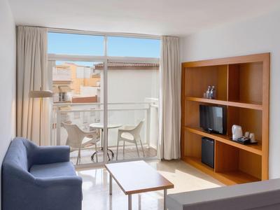 Helles Hotelzimmer mit Balkon, Sofa, Tisch und Regal mit Fernseher und Dekoration.
