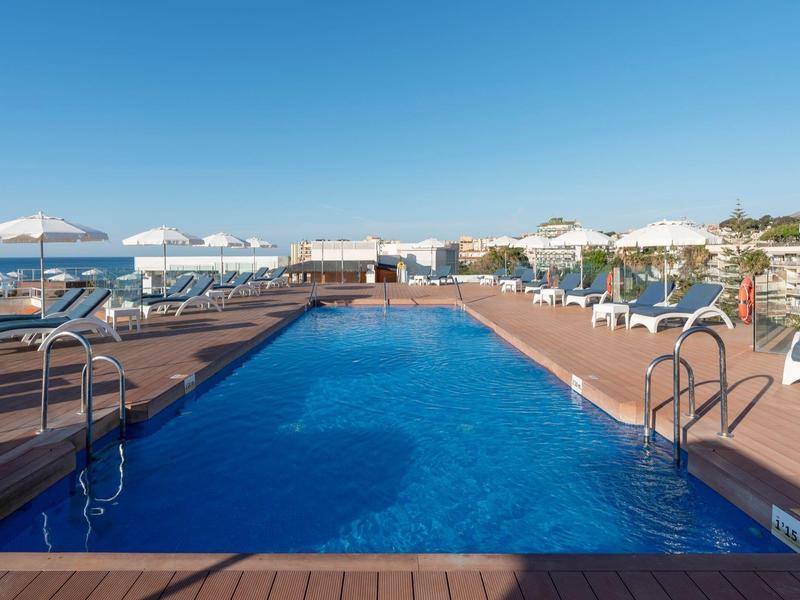 Outdoor hotel pool with blue water, surrounded by sun loungers and umbrellas under clear sky.
