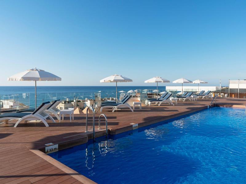 Outdoor pool with lounge chairs and umbrellas beside the sea under a clear sky.