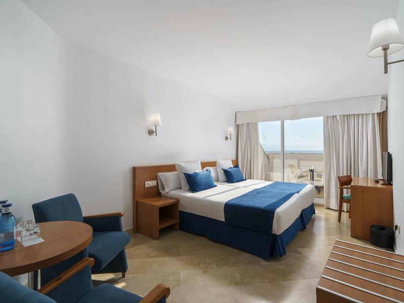 Bright hotel room with double bed, seating area, and balcony view of sea.