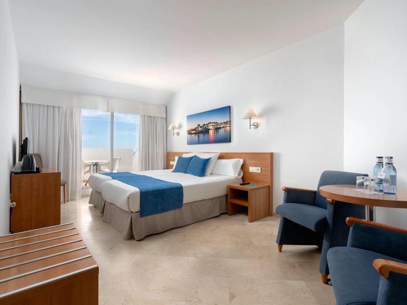Bright hotel room with double bed, blue accents, chairs, and city view through window.