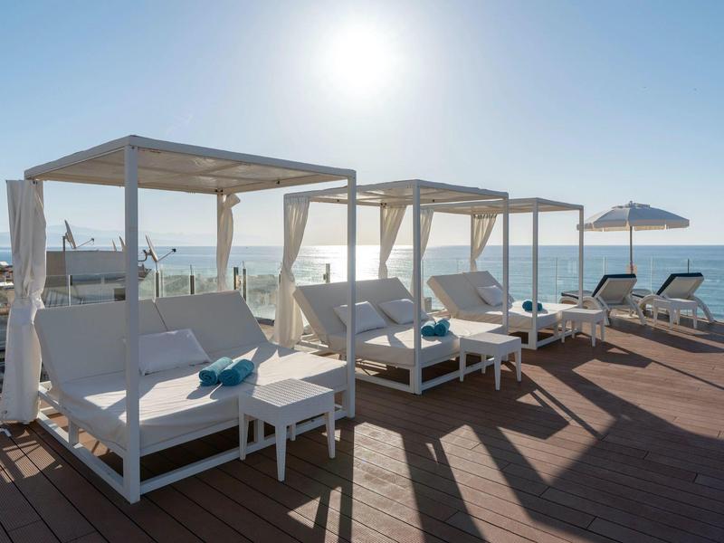 White canopy beds and lounge chairs on a wooden deck by the sea under a bright sun.