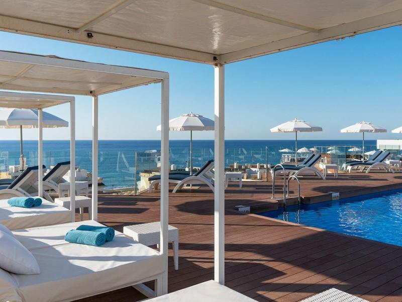 Poolside lounge beds with white umbrellas by the sea under a clear blue sky.