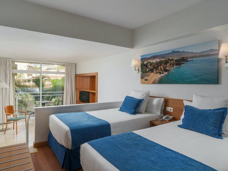 Hotel room with two beds, blue and white bedding, sea view painting, and a balcony with chairs.