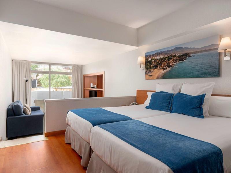 Bright hotel room with twin beds, blue accents, wooden floor, and a sea view painting.