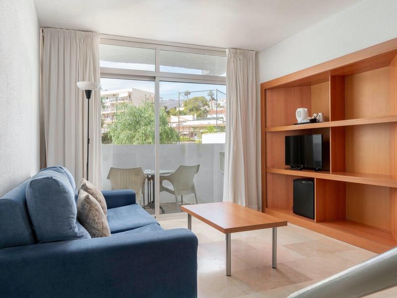 Cozy hotel room with a blue sofa, wooden shelves, balcony, and view outside.