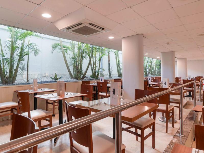 Spacious hotel dining area with wooden chairs and tables, large windows with greenery outside.