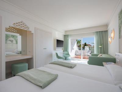 Hell eingerichtetes Hotelzimmer mit zwei Betten, Sitzbereich und Blick auf Balkon mit Meerblick.