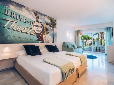 Helles Hotelzimmer mit großem Bett, Lounge-Sessel, dekorativer Wand und Blick auf Terrasse.