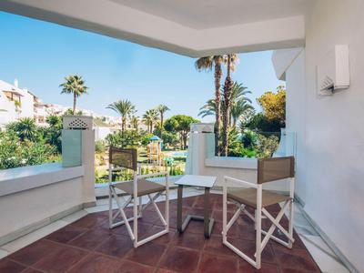 Balkon mit zwei Stühlen und kleinem Tisch, Blick auf Palmen und Ferienanlage bei klarem Himmel.