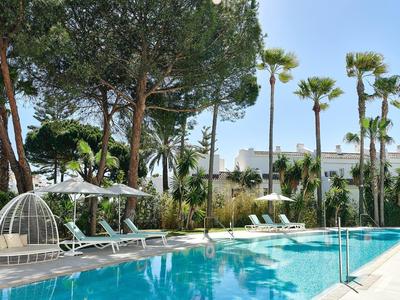 Pool mit Liegen und Hängesessel umgeben von Palmen und Hotelgebäude bei klarem Himmel.