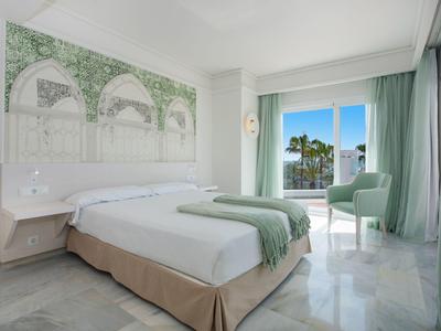 Helles Hotelzimmer mit Doppelbett, grün-weißer Wanddekoration und Balkon mit Meerblick.