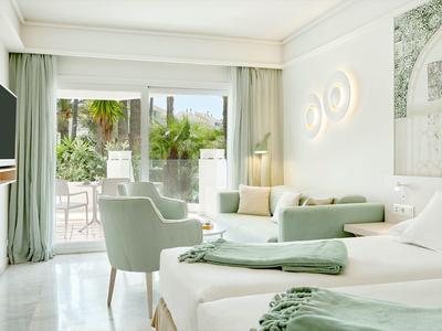 Helles Hotelzimmer mit zwei Betten, grünen Vorhängen und Zugang zu einer Terrasse mit Blick auf grüne Pflanzen.