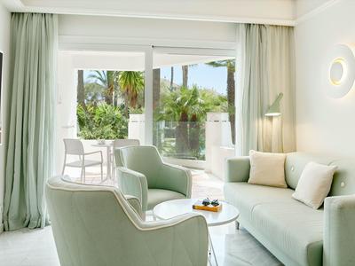 Helles Wohnzimmer mit grünen Sesseln, Sofa und Tür zu Balkon mit Blick auf Palmen.