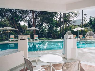 Piscina esterna moderna con area salotto, palme e ombrelloni in un ambiente tropicale dell'hotel.