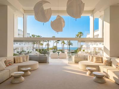 Helle Hotel-Lounge mit bequemen Sofas, runden Tischen und Blick auf Palmen und Meer.