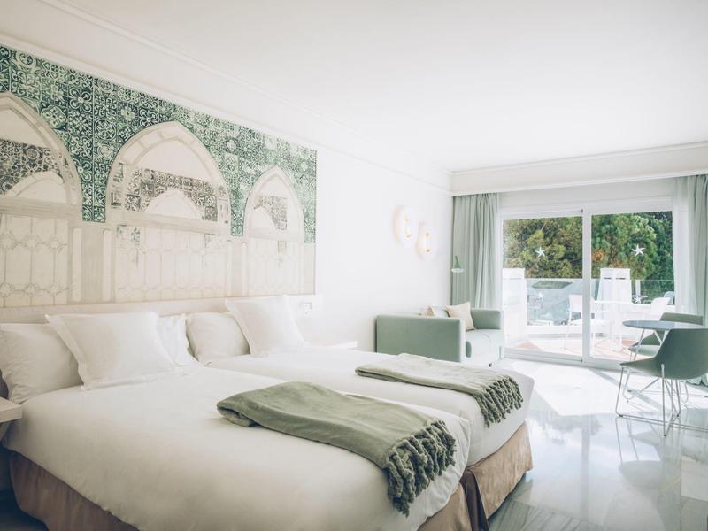 Bright hotel room with double bed, green wall art, and views of terrace with pool.