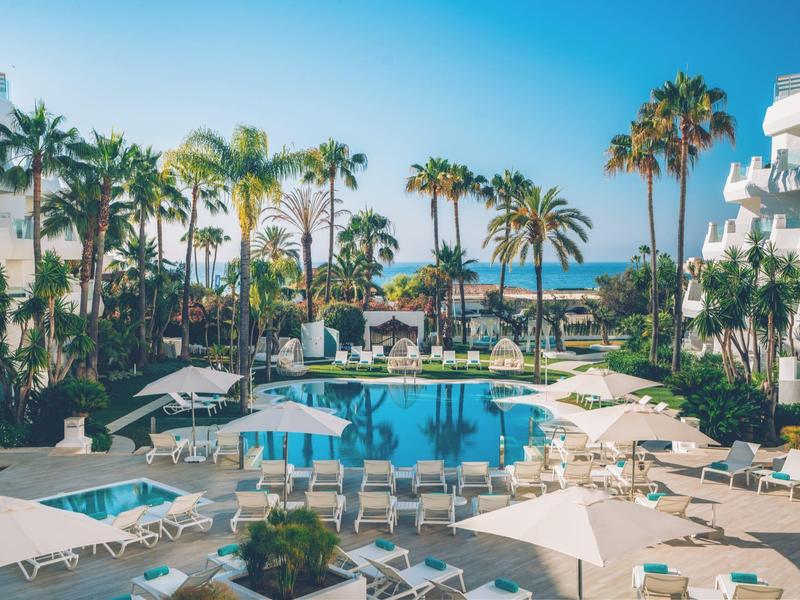 Luxury hotel pool with sun loungers, umbrellas, palm trees, blue sky, and ocean in the background.